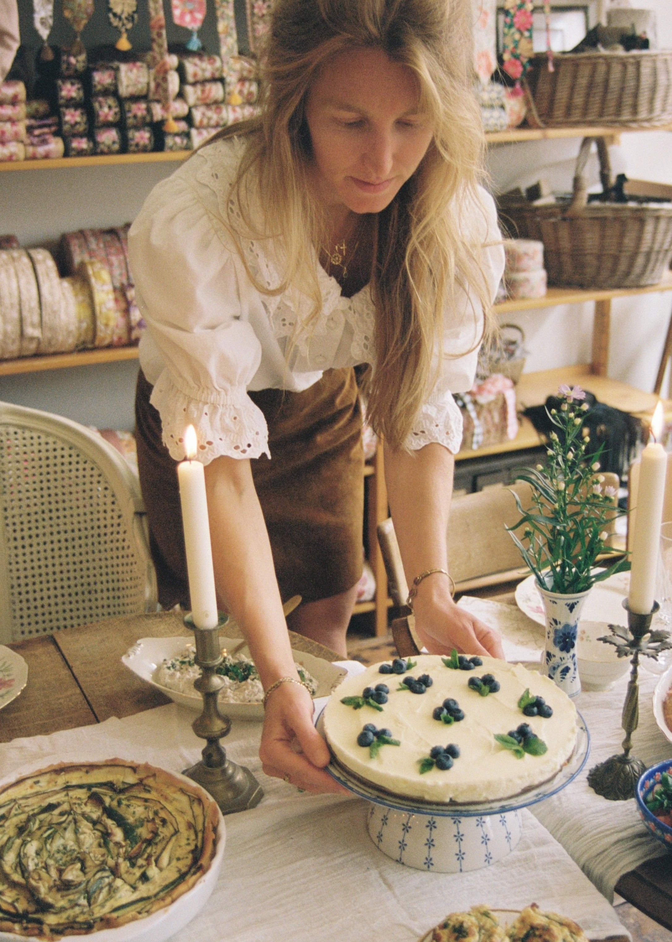 Vrouw in een romantische blouse serveert een taart met blauwe bessen aan een sfeervol gedekte tafel met kaarslicht en bloemen in een ambachtelijke setting.