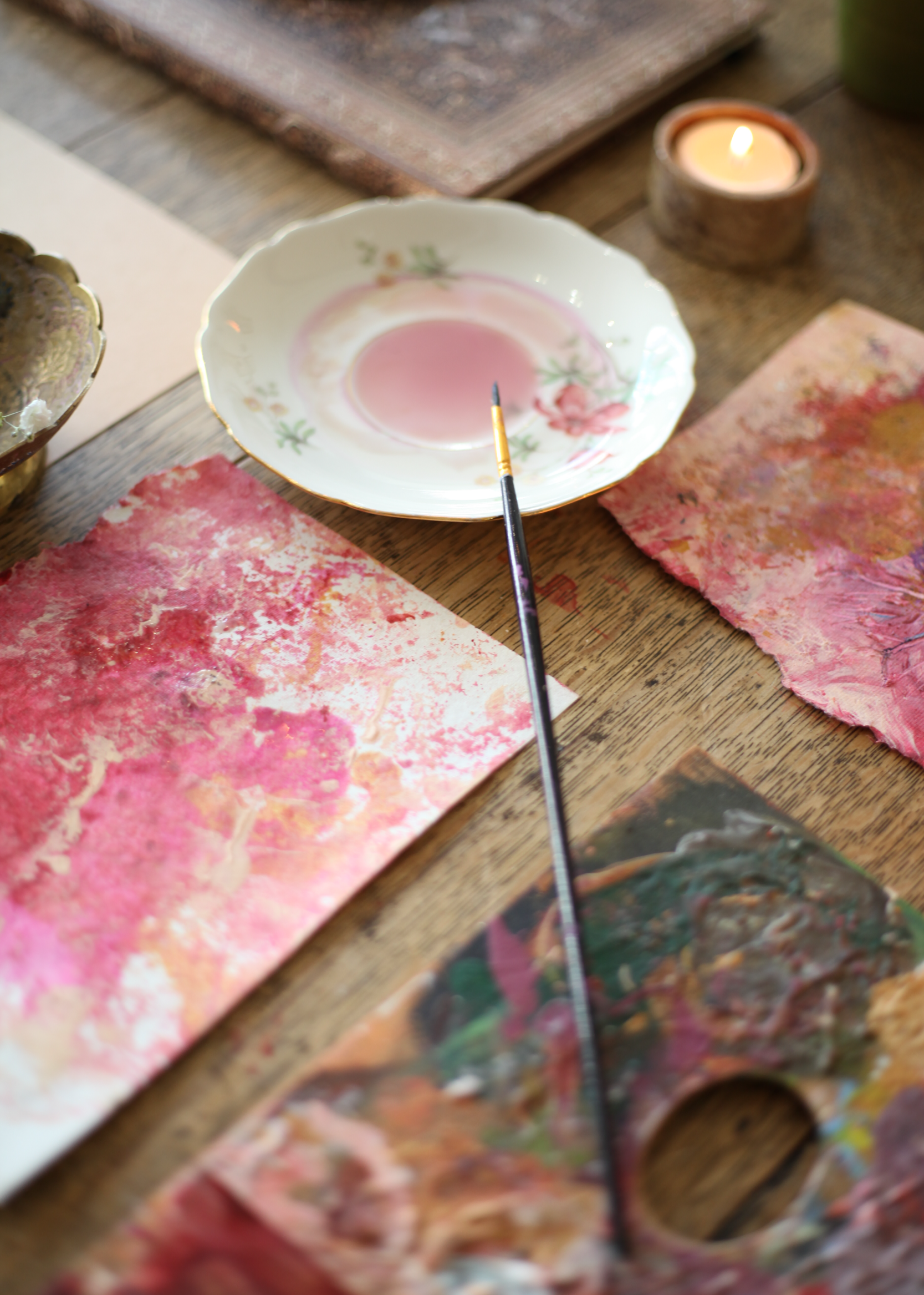 Creatieve schilderworkshop met roze aquarelverf, een penseel en handgeschept papier op een rustieke houten tafel, omringd door sfeervol kaarslicht.