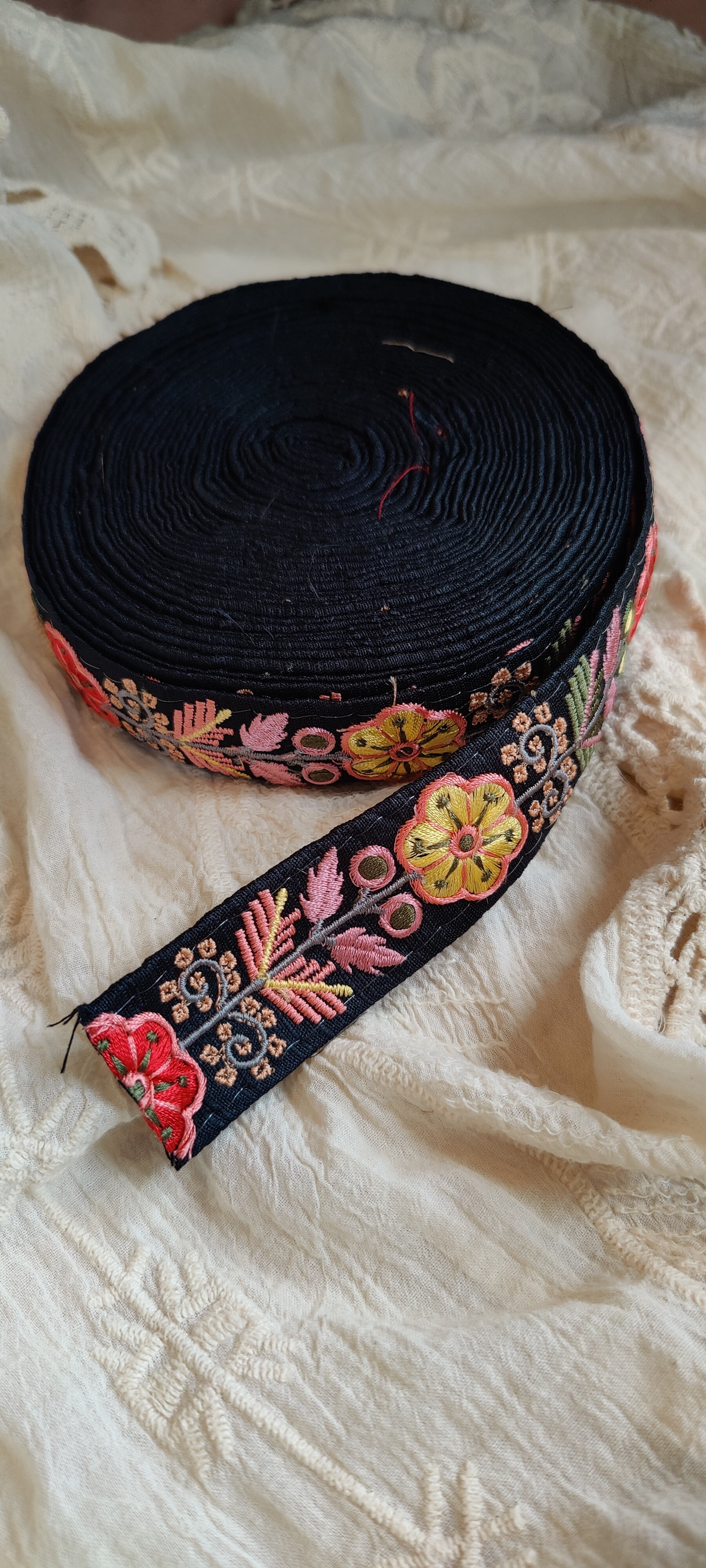 Roll of black ribbon with colorful floral embroidery on a textured white fabric background